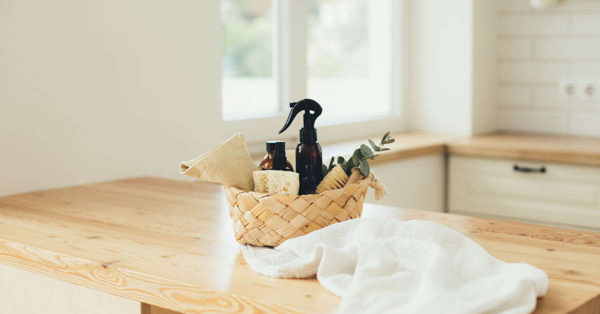 Zero waste siivoustarvikkeet korissa puupöydällä: suihkepullo, pesusieni ja liina ekologiseen siivoukseen vaaleassa keittiössä.
